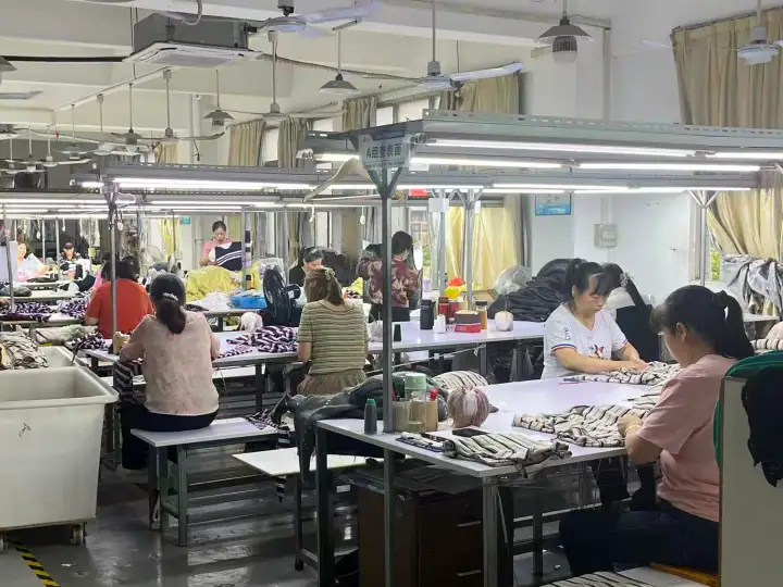 Workers in a clothing manufacturing workshop sewing and assembling garments at workstations with bright lighting and industrial equipment.