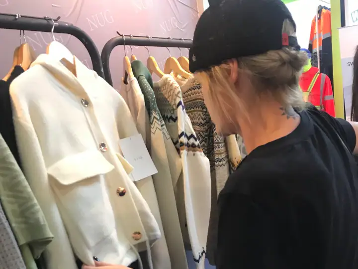 Young woman shopping for stylish sweaters and coats at NUBIG Fashion store, browsing a clothing rack with various knitwear and outerwear.