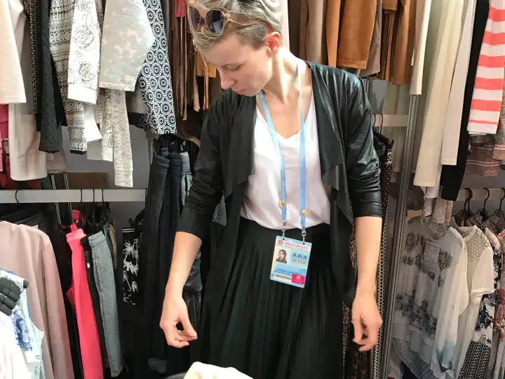 Female retail worker browsing clothing racks in a fashion store, wearing a black jacket and white top, with a store badge around her neck.