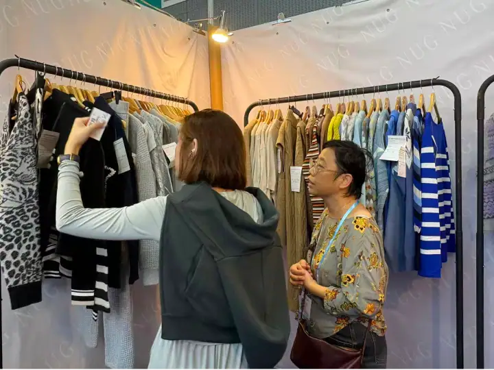 Two women shopping for clothes at a local market, browsing through racks of various garments including sweaters and striped shirts.
