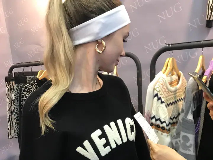 Young woman with blonde hair, white headband, and gold hoop earrings shopping for clothes at a retail store, holding a smartphone and looking at a clothing tag.