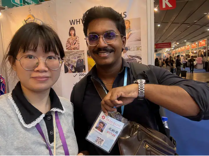Two diverse attendees at a fashion trade show, smiling and showing their badges, with booths and other visitors in the background.