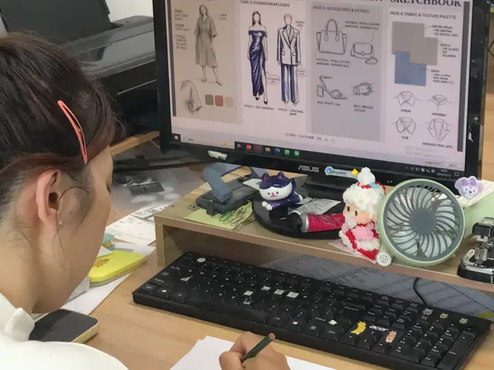 A person working on fashion design sketches on a computer screen, surrounded by plush toys and design tools, in a creative workspace.