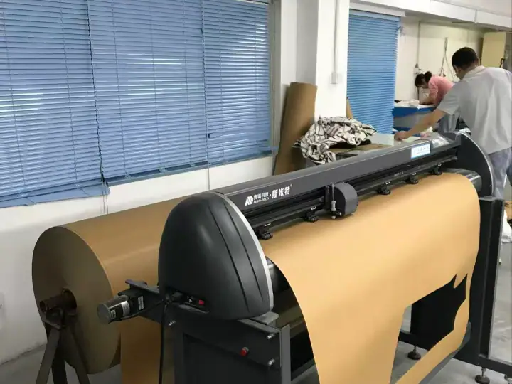 Industrial large format printer printing on fabric roll in a factory setting with workers in the background.