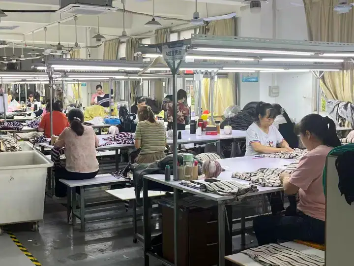 Workers in a textile factory sewing and assembling fabrics under bright lighting, with multiple workstations and sewing machines.