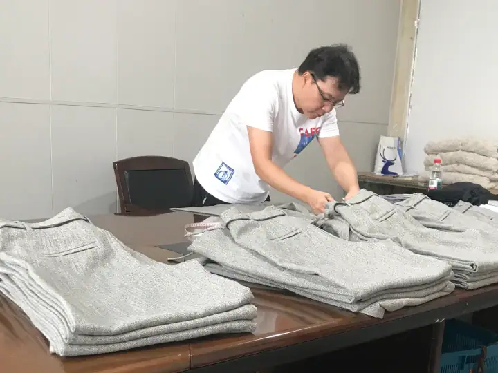 Man folding grey sweaters on a table in a clothing store or warehouse setting, with stacks of similar sweaters around him.