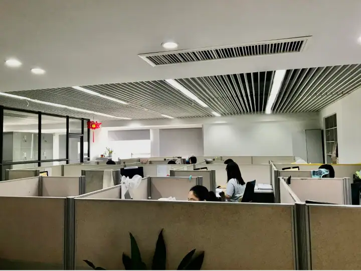 Open-plan office with beige cubicles, employees working at desks, and a sleek ceiling design with linear lighting.