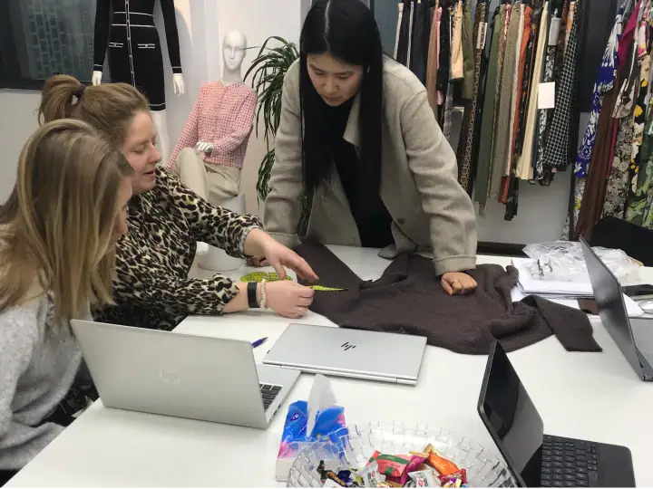 Three women collaborating on fashion design in a boutique, discussing fabric and style options with laptops and fabric samples.