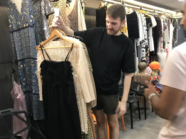 Man shopping for elegant dresses at a clothing boutique, browsing through a rack of stylish garments with a smile.