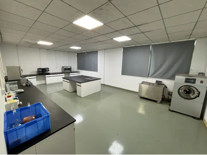 Spacious laundry room with washing machines, dryers, and work tables, featuring large windows with blinds and a clean, organized environment.
