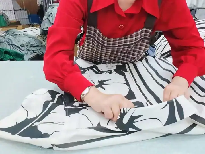 Person in red shirt and checkered apron cutting fabric with black and white abstract pattern in a textile workshop.