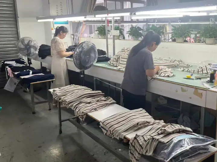 Workers inspecting and folding striped fabric in a textile factory, with large fans and potted plants in the background.
