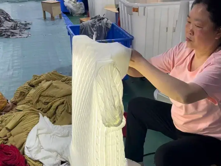 Worker inspecting and handling fabric in a clothing manufacturing facility, with piles of textiles and industrial equipment in the background.