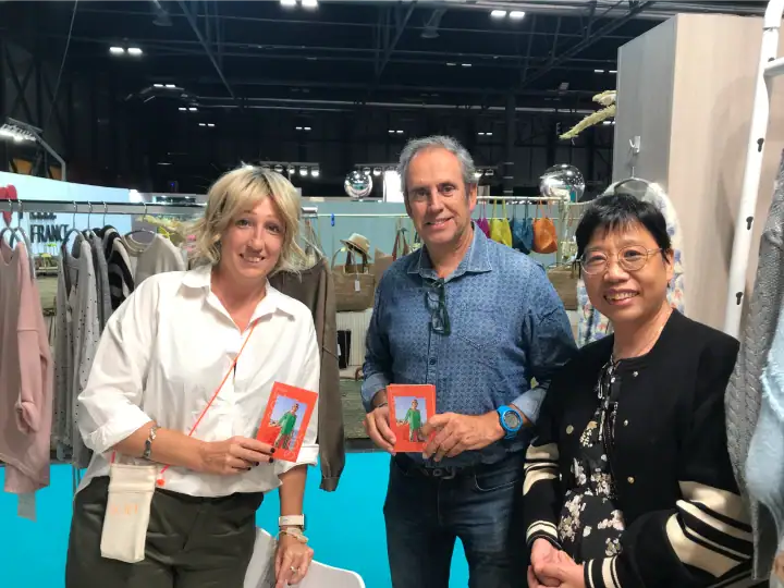 Three people at a fashion event holding small red and green items, smiling, with clothing displays in the background.
