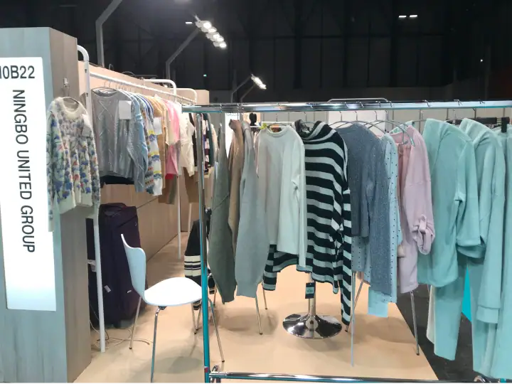 Clothing display at NINGBO United Group booth showcasing sweaters, shirts, and casual wear in a trade show setting.