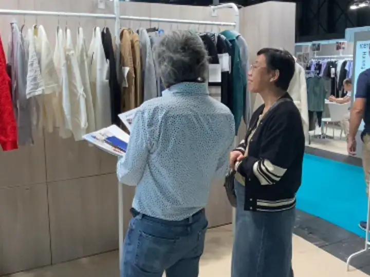Two women engaged in conversation at a clothing trade show, with racks of garments in the background.