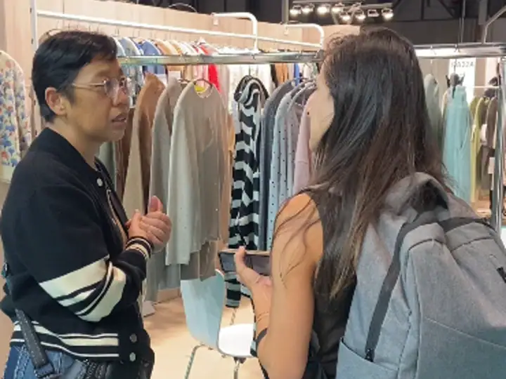 Two women engaging in a conversation inside a clothing store, with racks of colorful garments in the background.