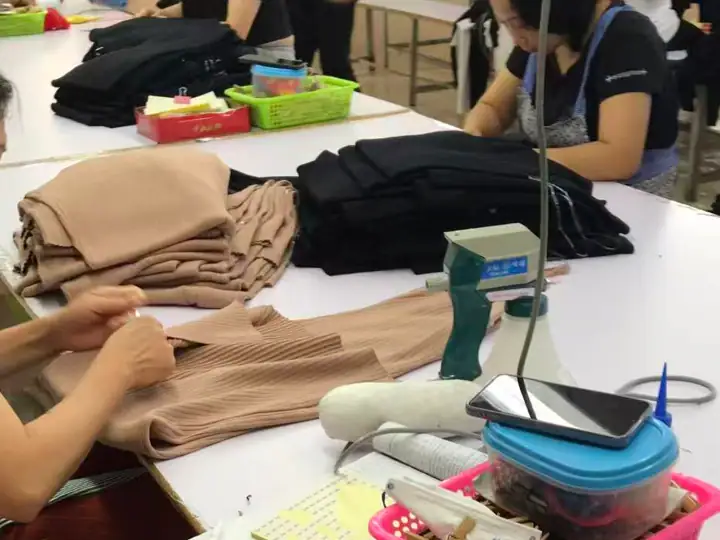 Workers sorting and folding clothing in a manufacturing facility with various fabric pieces and tools on the table.