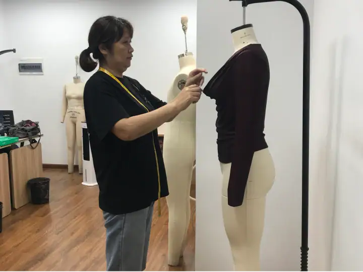 Fashion designer working on a dark-colored long-sleeve top on a mannequin in a studio setting, with sewing tools and additional mannequins in the background.
