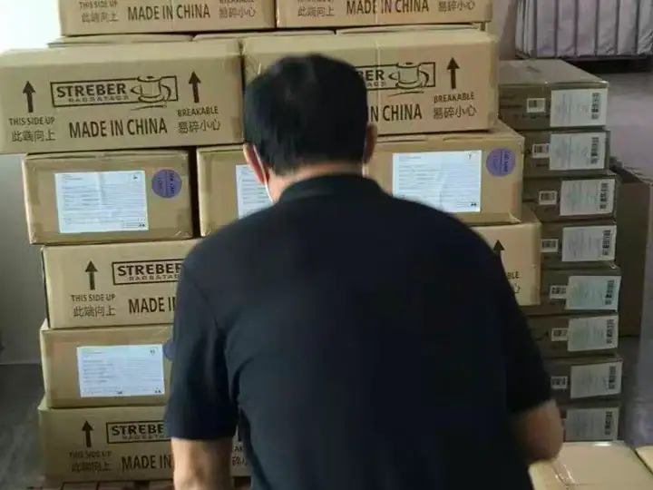 A warehouse worker with black hair, wearing a black shirt, is stacking or inspecting cardboard boxes labeled 'Made in China' in a storage facility.