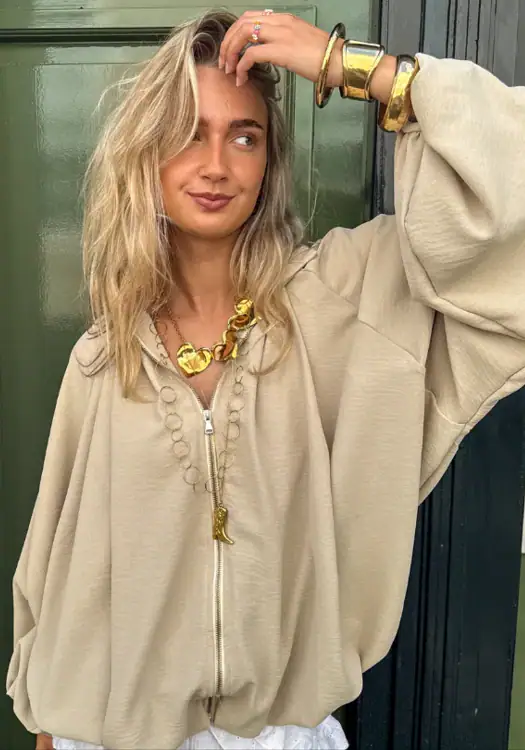 Stylish woman wearing a beige oversized zip-up jacket and bold gold jewelry including a chunky necklace and bracelets, posing outdoors.