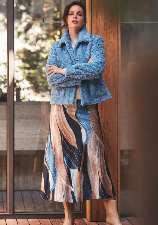 Fashionable woman wearing a blue fuzzy coat and a colorful patterned skirt, standing outdoors near a wooden door with glass panels.