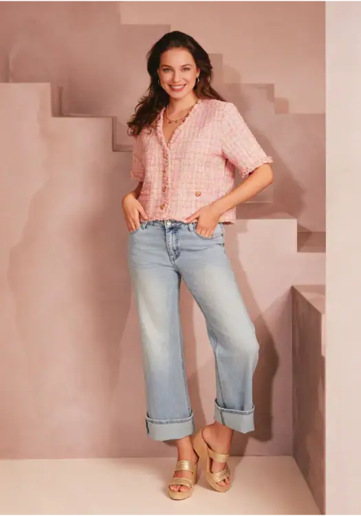 Young woman in pink tweed jacket and light blue jeans, smiling, standing indoors with stairs in the background.