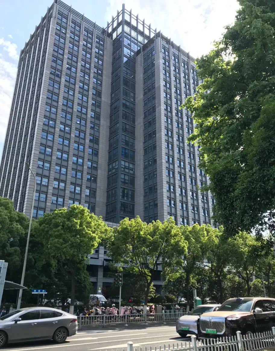Tall modern office skyscraper with glass facade surrounded by green trees and busy street scene in an urban environment.
