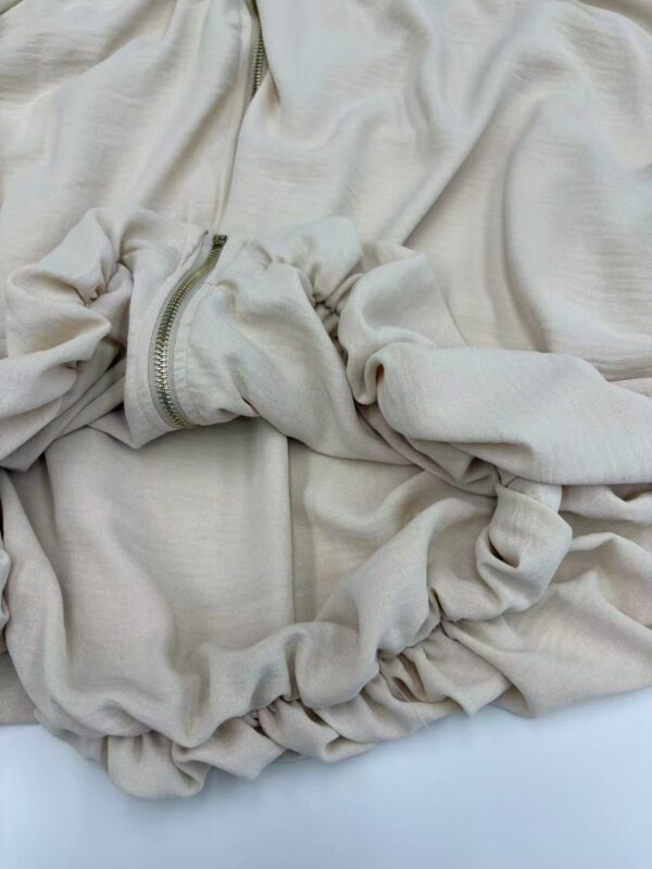 Close-up of a cream-colored zip-up hoodie with a front pocket and drawstrings, made from soft fabric, laid flat on a white surface.