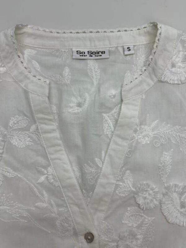 Close-up of a white embroidered blouse with floral patterns and a small button at the neckline, perfect for stylish summer wear.