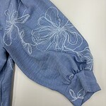 Close-up of a blue blouse with white floral embroidery on the sleeve, showcasing elegant design and craftsmanship.