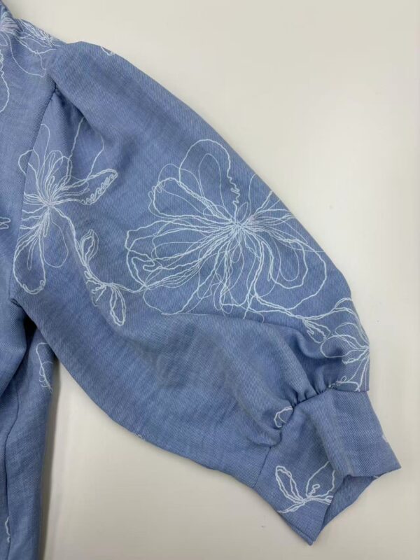 Close-up of a blue blouse with white floral embroidery on the sleeve, showcasing elegant design and craftsmanship.