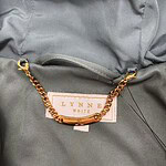 Close-up of a gray jacket featuring a gold chain accessory attached to a label that reads 'LYNNB WHITE'.