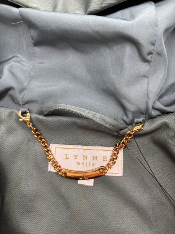 Close-up of a gray jacket featuring a gold chain accessory attached to a label that reads 'LYNNB WHITE'.