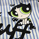 Adorable cartoon character with large green eyes, black hair, and a playful expression, sitting on blue and white striped fabric with small black bows.