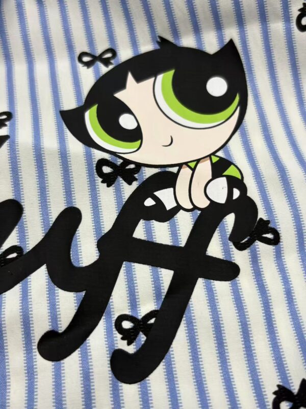 Adorable cartoon character with large green eyes, black hair, and a playful expression, sitting on blue and white striped fabric with small black bows.