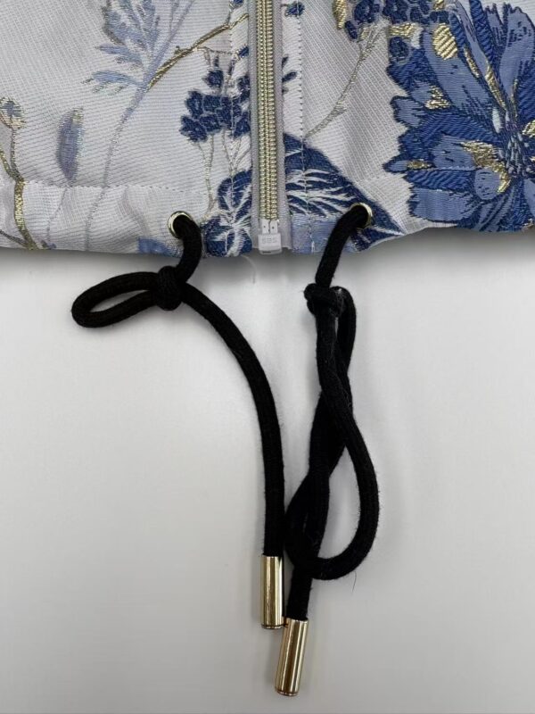 Close-up of a floral patterned jacket featuring a zipper and black drawstrings with gold tips, showcasing detailed embroidery and fabric texture.