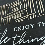 Close-up of fabric with stylish text design, featuring the phrase 'Enjoy the little things' in elegant script and modern font, ideal for fashion or home decor.