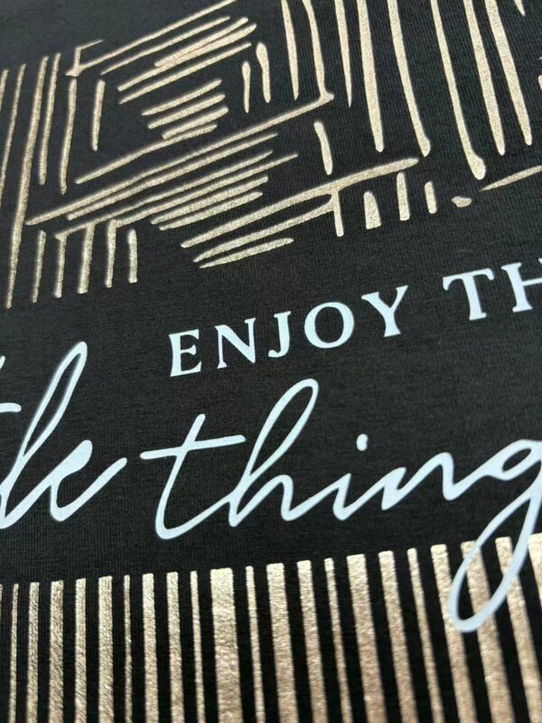 Close-up of fabric with stylish text design, featuring the phrase 'Enjoy the little things' in elegant script and modern font, ideal for fashion or home decor.