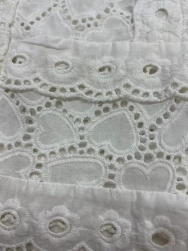 Close-up of white embroidered fabric with intricate eyelet and floral patterns, showcasing delicate craftsmanship and fine detailing.