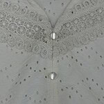 Close-up of a white dress featuring intricate lace embroidery, pearl buttons, and eyelet fabric, highlighting craftsmanship and delicate design.