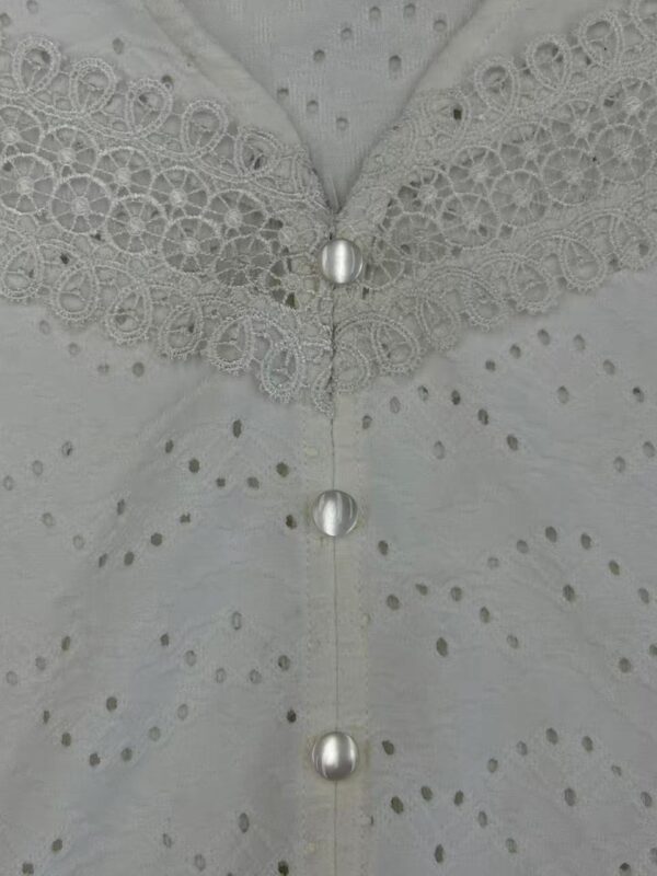 Close-up of a white dress featuring intricate lace embroidery, pearl buttons, and eyelet fabric, highlighting craftsmanship and delicate design.