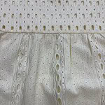 Close-up of white eyelet fabric with intricate cut-out patterns and embroidery, showcasing delicate craftsmanship and lightweight material suitable for summer fashion.