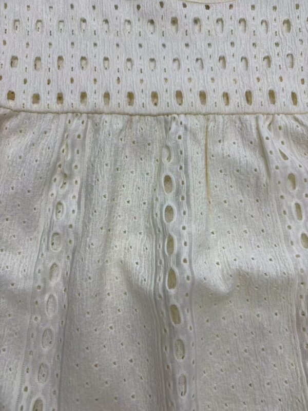 Close-up of white eyelet fabric with intricate cut-out patterns and embroidery, showcasing delicate craftsmanship and lightweight material suitable for summer fashion.