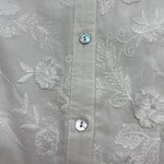 Close-up of a white shirt with intricate floral embroidery and shiny buttons, showcasing detailed craftsmanship and elegant design.