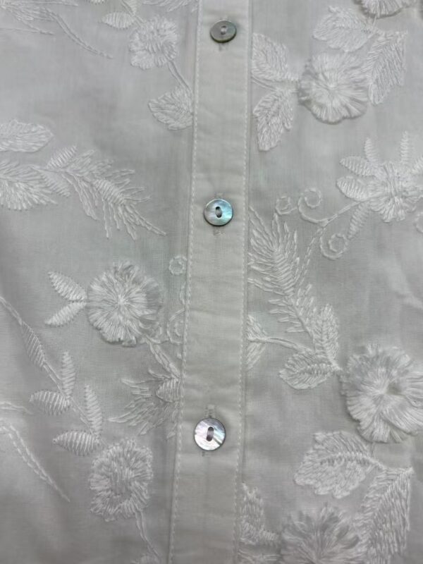 Close-up of a white shirt with intricate floral embroidery and shiny buttons, showcasing detailed craftsmanship and elegant design.