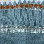 Close-up of a knitted sweater with geometric patterns in blue, orange, and white, showcasing detailed knitting and vibrant colors.
