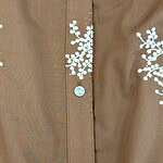 Close-up of a brown shirt featuring white floral embroidery and light blue buttons, showcasing detailed craftsmanship and stylish design.