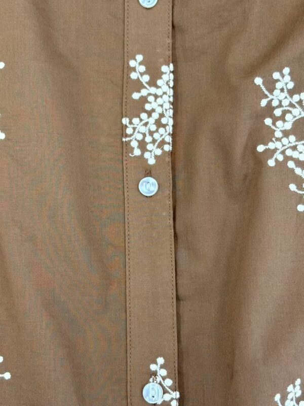 Close-up of a brown shirt featuring white floral embroidery and light blue buttons, showcasing detailed craftsmanship and stylish design.