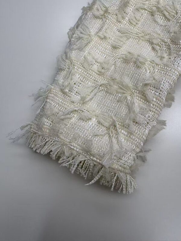 Close-up of a cream-colored textured fabric with intricate woven patterns and fringed edges, ideal for fashion or home decor.